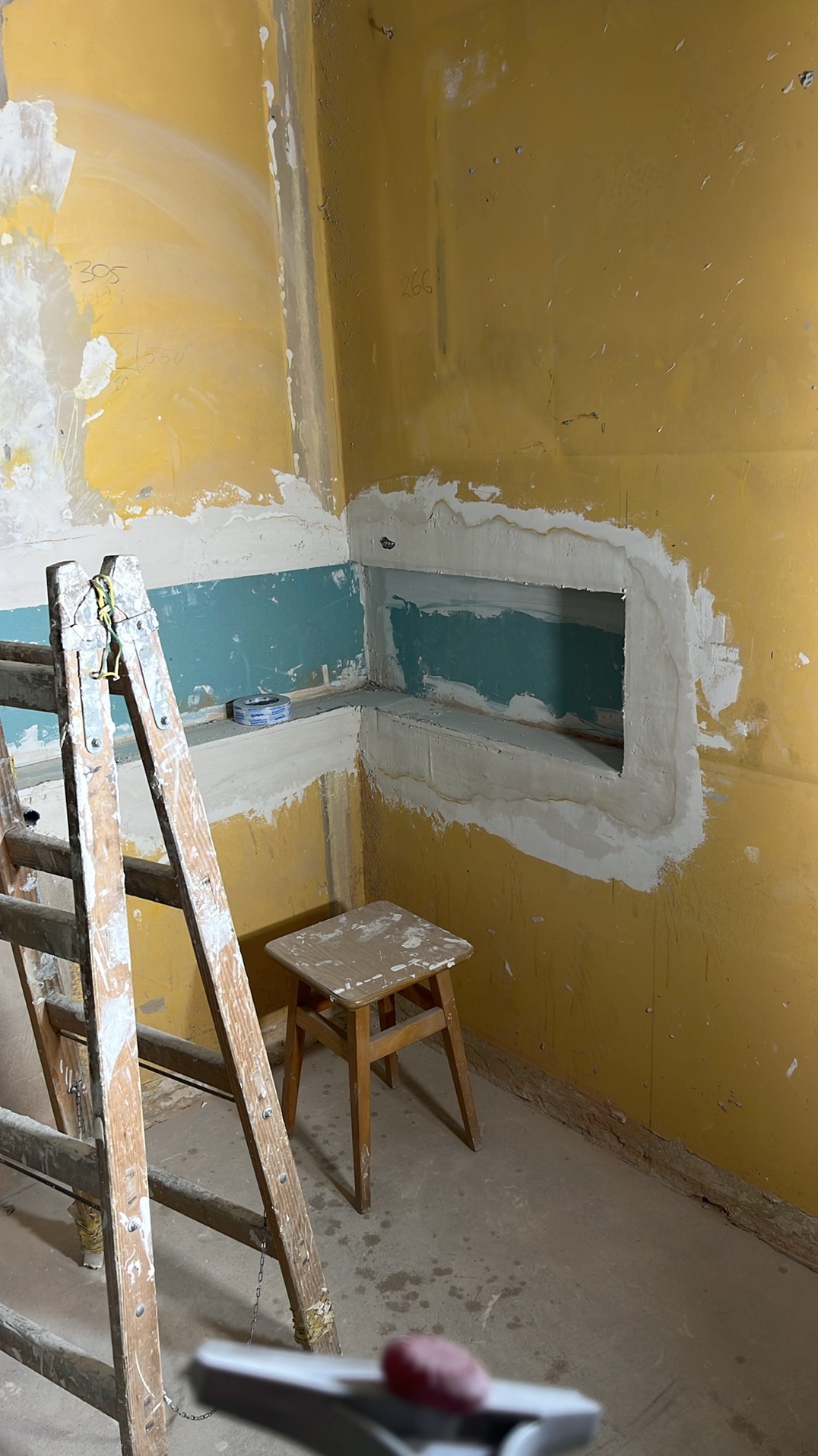 Żółta ściana z wyciętym otworem, przygotowana do szpachlowania, widoczne ślady po wcześniejszych warstwach, drabina malarska i stołek w pomieszczeniu w trakcie remontu.