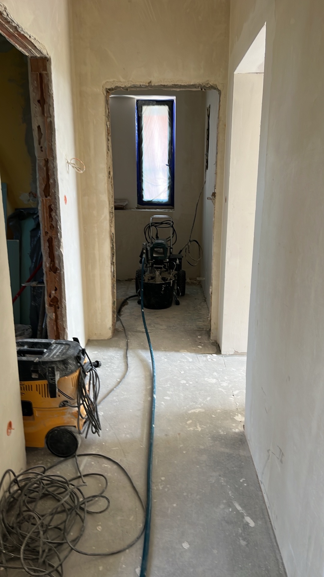 Wnętrze w trakcie prac wykończeniowych: ściany po nałożeniu gładzi, okno zabezpieczone folią, agregat malarski i odkurzacz przemysłowy na podłodze.