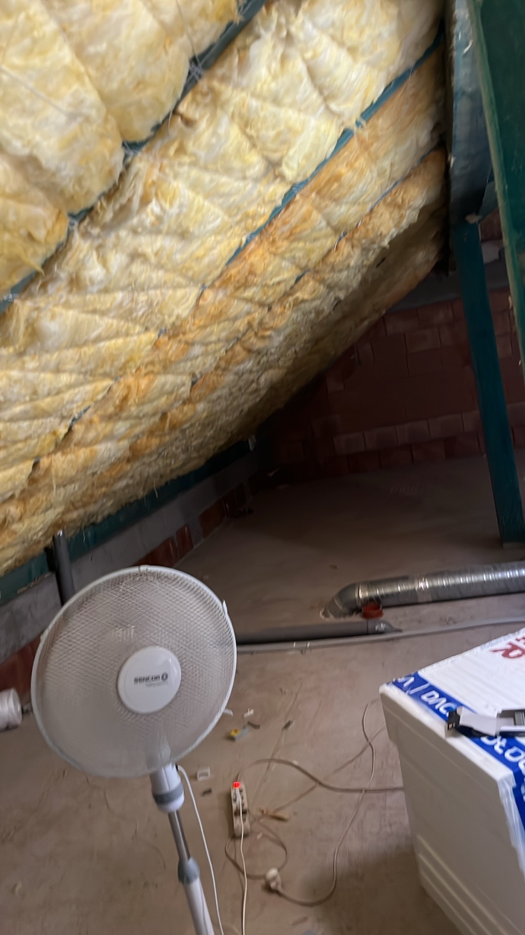 Ocieplenie poddasza wełną mineralną z widoczną konstrukcją dachu, wentylatorem Sencor i styropianem w tle.