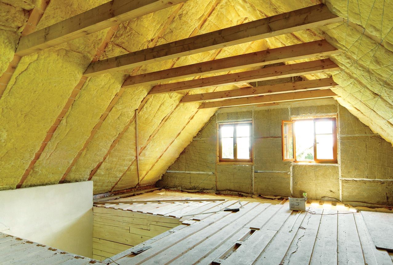 Poddasze w trakcie ocieplania wełną mineralną, widoczne drewniane belki konstrukcyjne, dwa okna z drewnianą stolarką i częściowo ułożona drewniana podłoga.