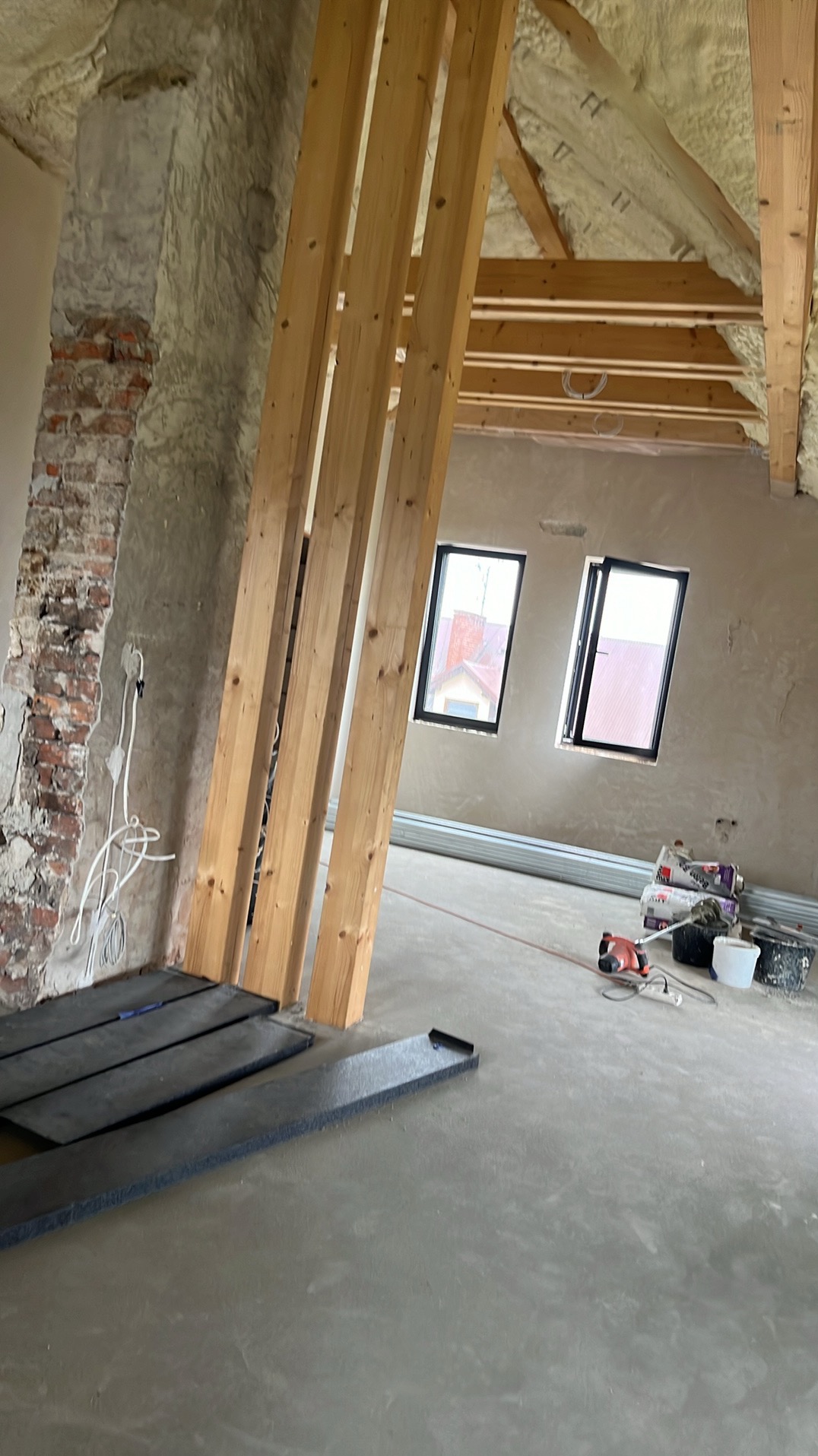 Poddasze w trakcie prac wykończeniowych: drewniane belki konstrukcyjne, ocieplenie pianką, przygotowane płyty izolacyjne na podłodze, widoczne okna dachowe i narzędzia pracy.
