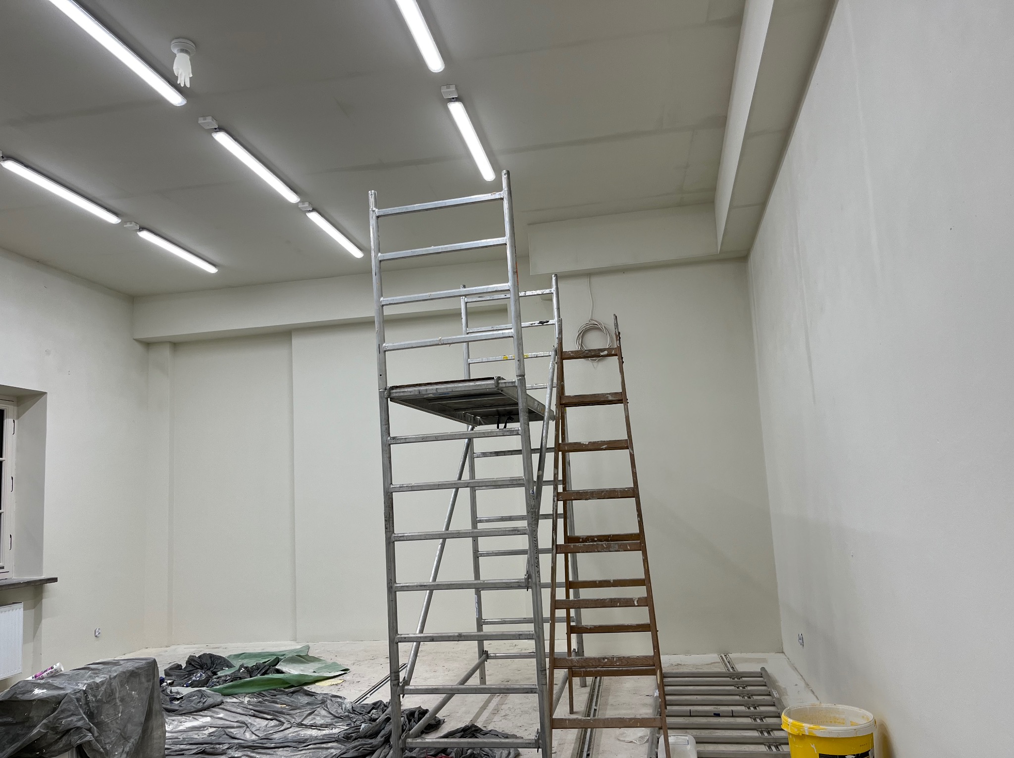 Wnętrze w trakcie prac wykończeniowych: świeżo pomalowane ściany na biało, dwa rodzaje drabin (aluminiowa i drewniana), zabezpieczona podłoga folią malarską, widoczne lampy sufitowe.