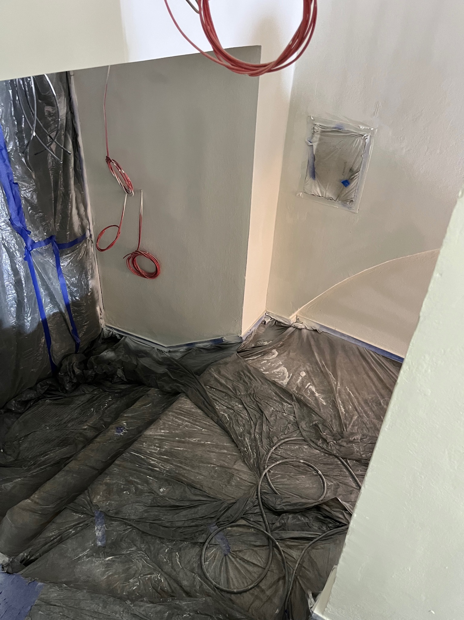 Przygotowane pomieszczenie do prac malarskich: podłoga i fragment ściany zabezpieczone folią i taśmą malarską, widoczne kable elektryczne.