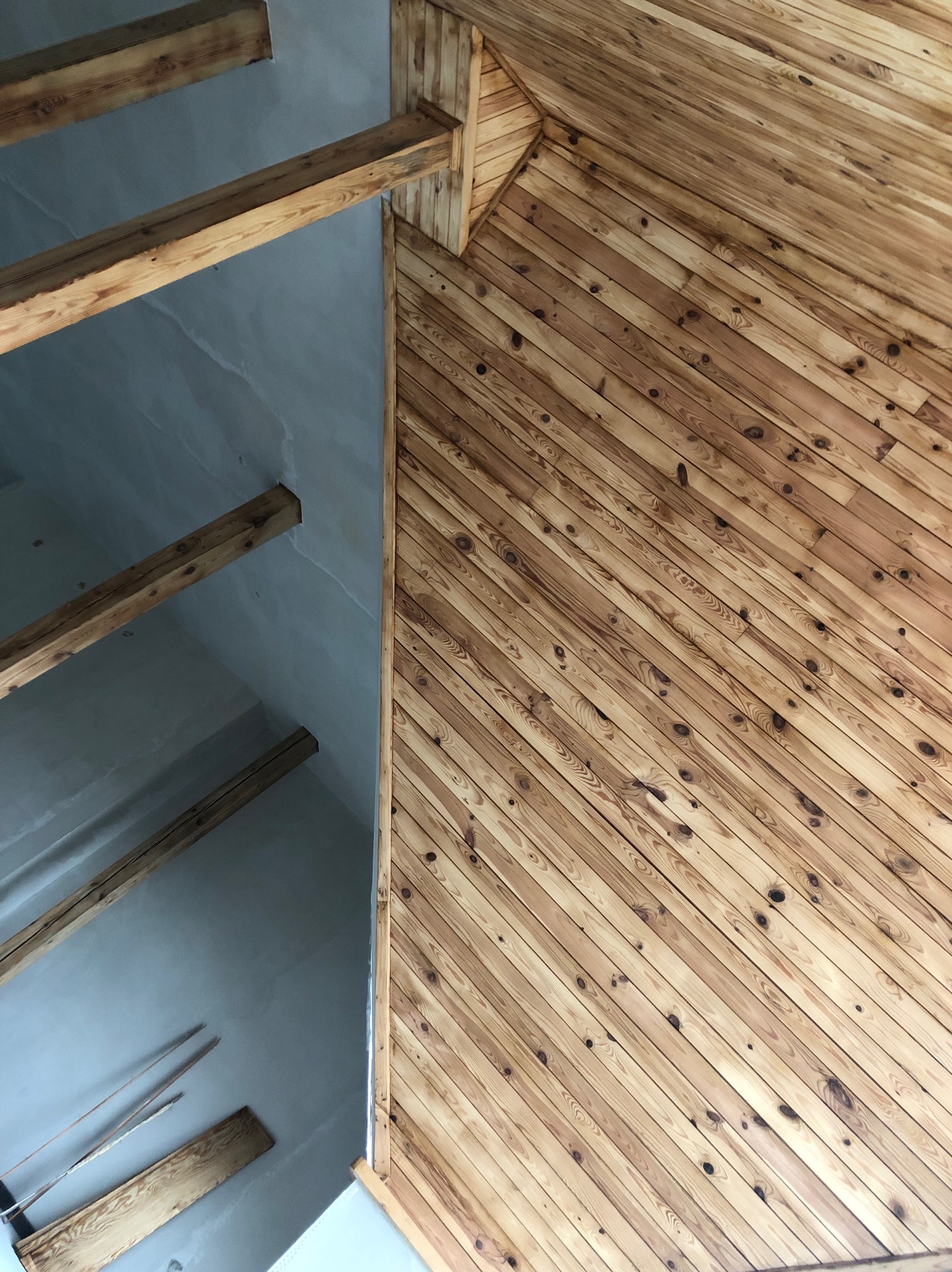 Skośny sufit wykończony drewnianą boazerią w jasnym odcieniu, kontrastujący z fragmentem surowego, pomalowanego na szaro stropu z widocznymi drewnianymi belkami.