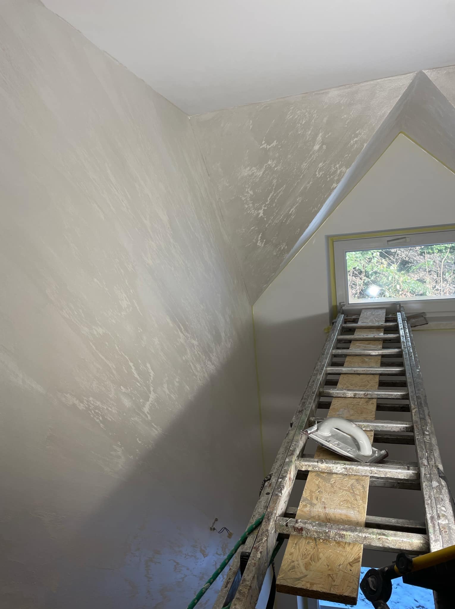 Ściana poddasza po nałożeniu dekoracyjnej, szarej gładzi, widoczna drabina oparta o okno z narzędziem do gładzi na szczeblu.