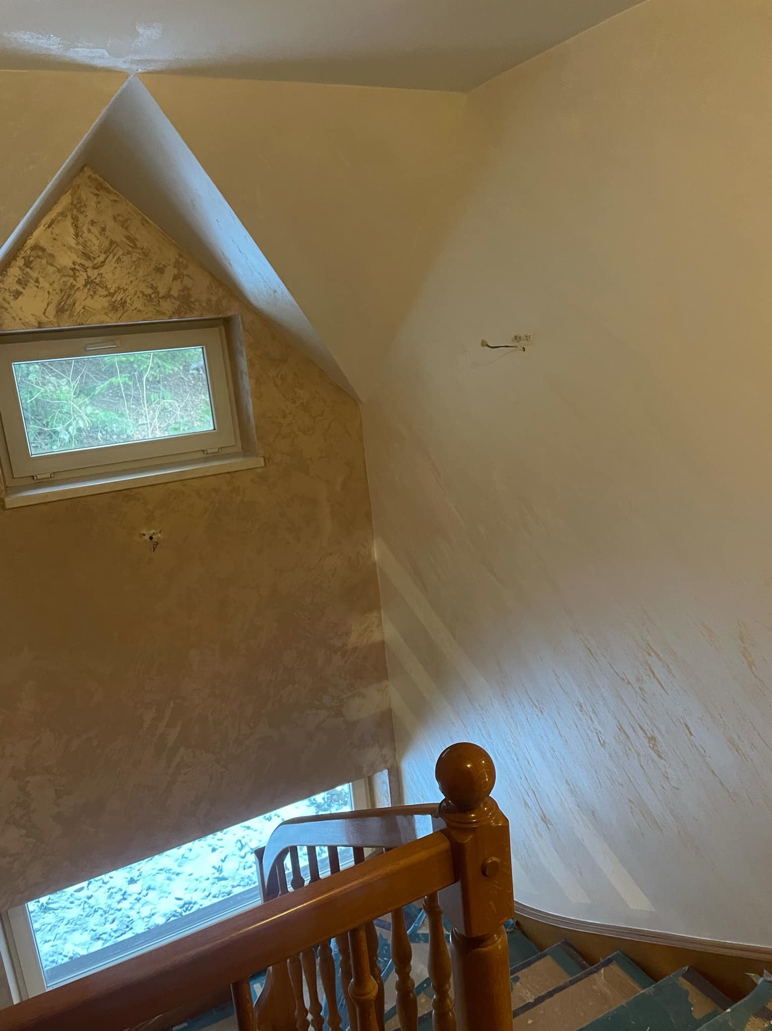 Klatka schodowa w domu z wykończonymi ścianami w odcieniach beżu i brązu, jedna ze ścian z efektem dekoracyjnym, widoczne schody zabezpieczone folią.