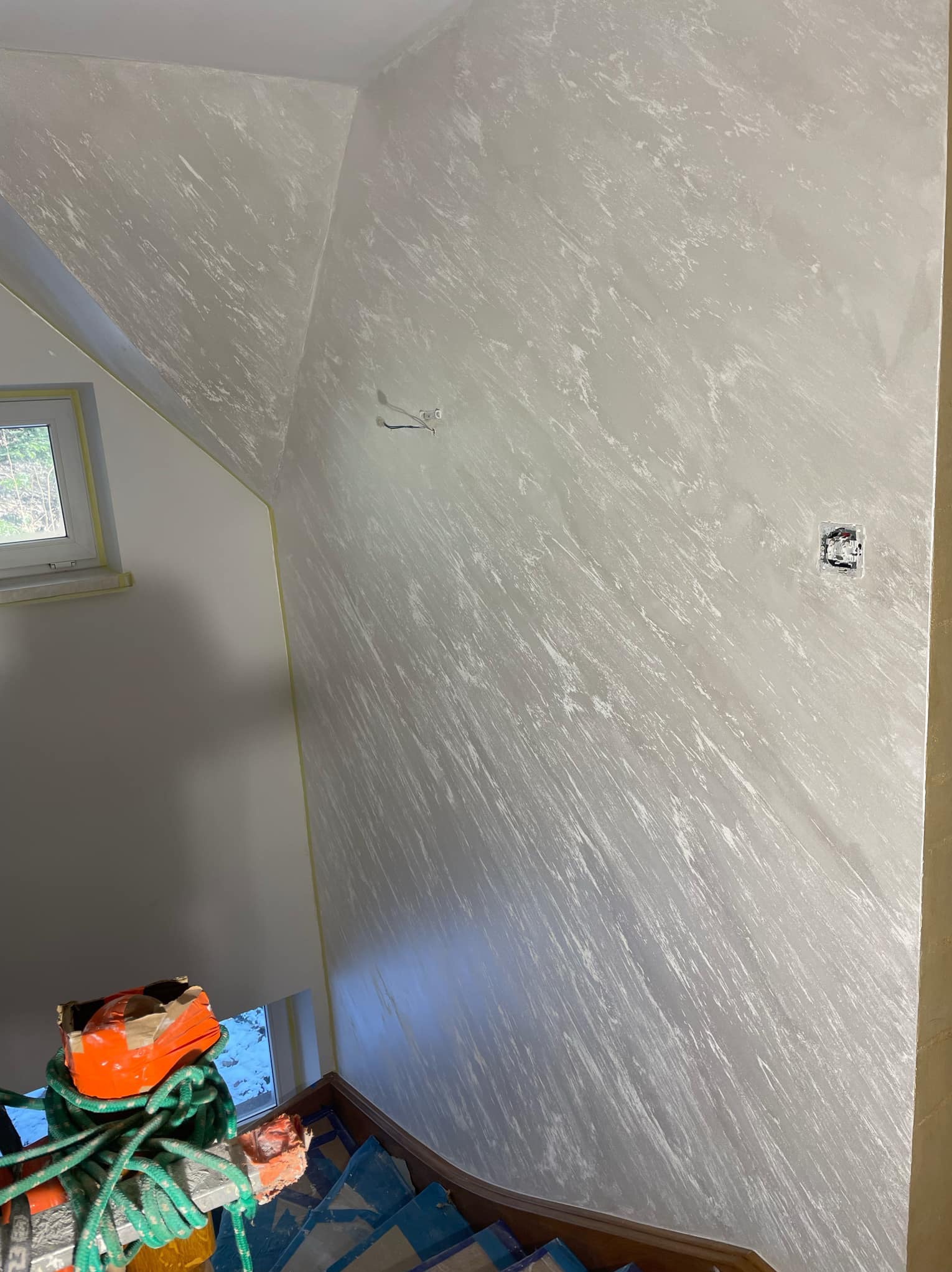 Ściana wykończona dekoracyjną gładzią w odcieniach szarości z widoczną fakturą, zabezpieczona taśmą malarską przy oknie i schodach, z widocznymi puszkami elektrycznymi.