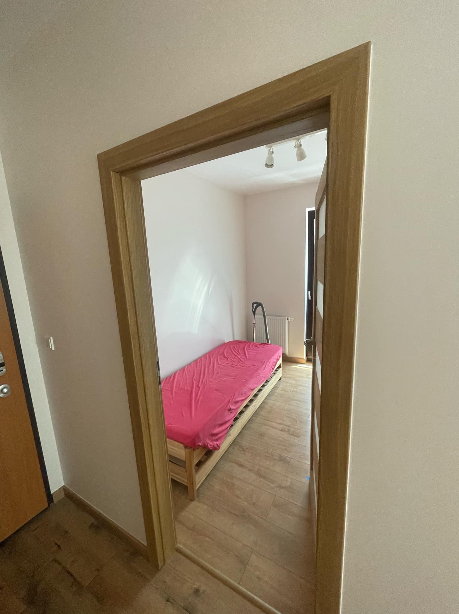 Widok przez drewnianą futrynę na pokój z łóżkiem z różową pościelą, odkurzaczem i grzejnikiem przy ścianie, widoczne fragmenty podłogi z paneli.