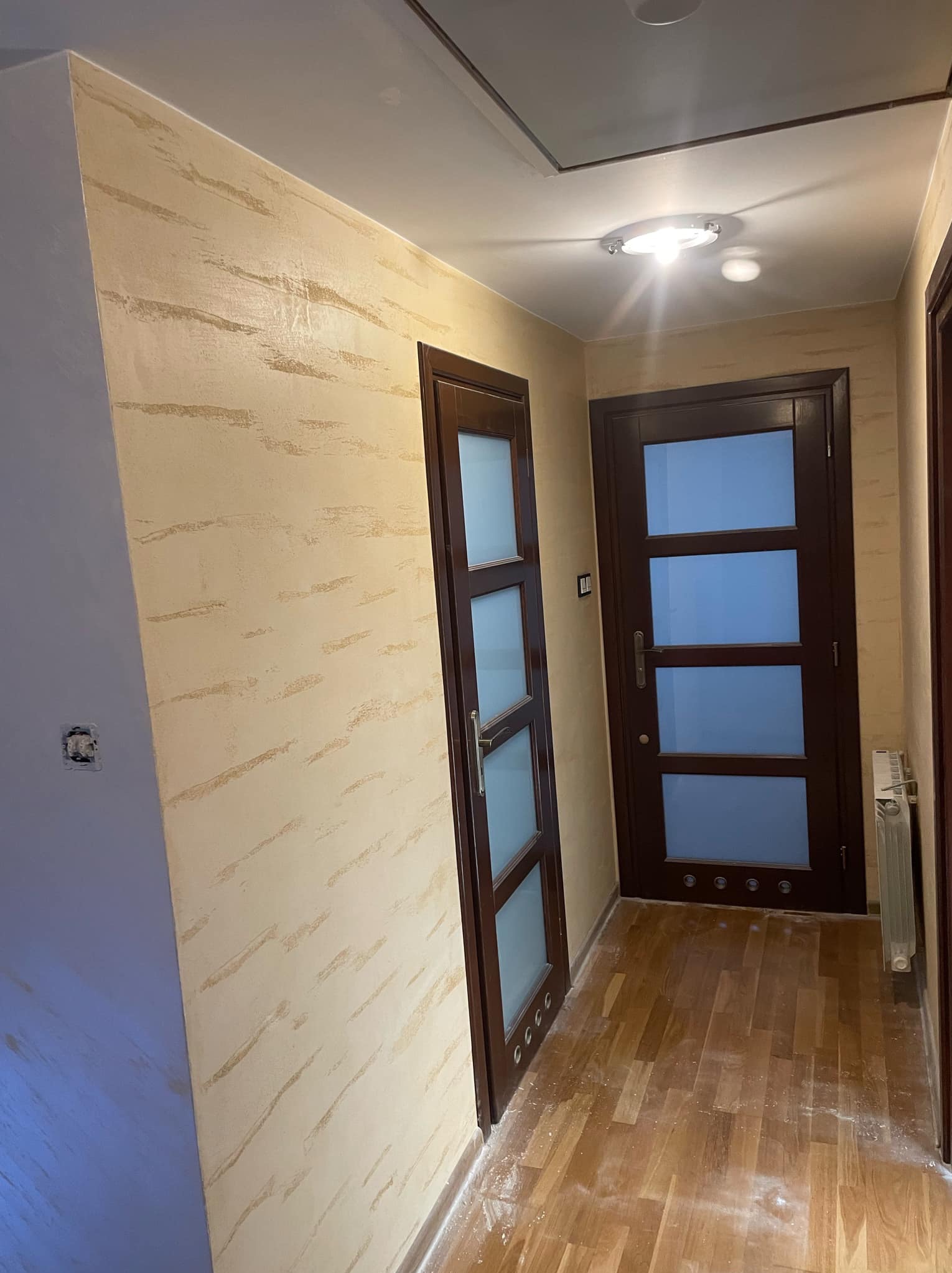 Korytarz po remoncie z dekoracyjną, beżową ścianą o fakturze przypominającej piasek, drewnianą podłogą i dwiema ciemnymi, przeszklonymi drzwiami.