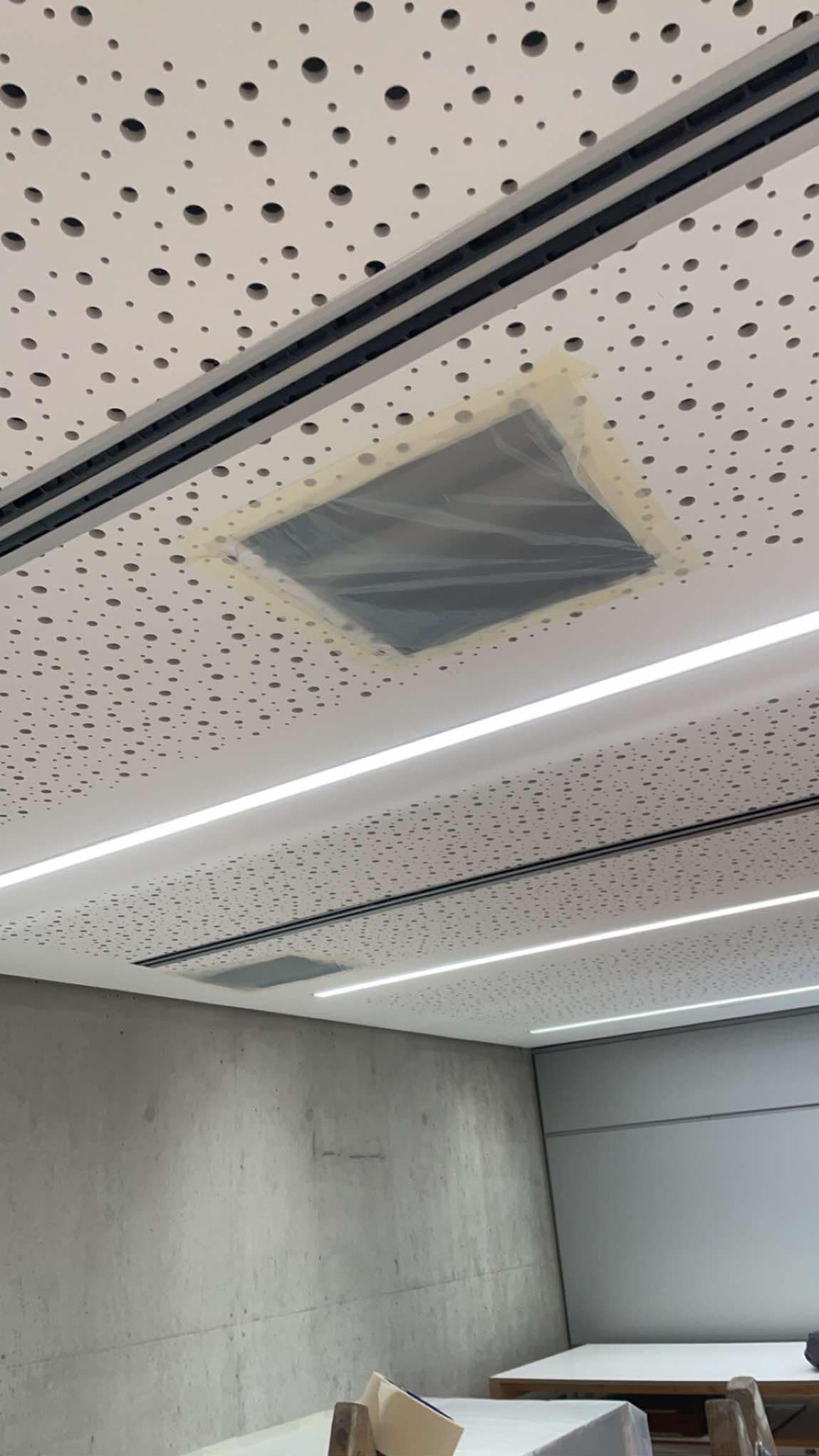 Montaż sufitu podwieszanego z perforowanymi płytami, zabezpieczone otwory na oświetlenie taśmą malarską i folią, widoczne ledowe listwy oświetleniowe.