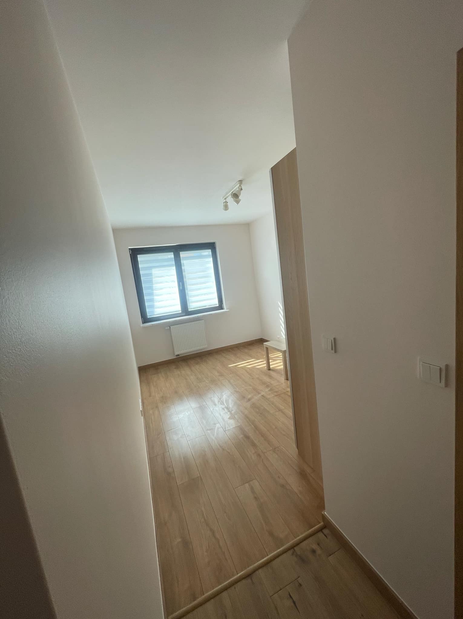 Widok z korytarza na pokój po remoncie: świeżo położone panele podłogowe, białe ściany i okno z żaluzjami.