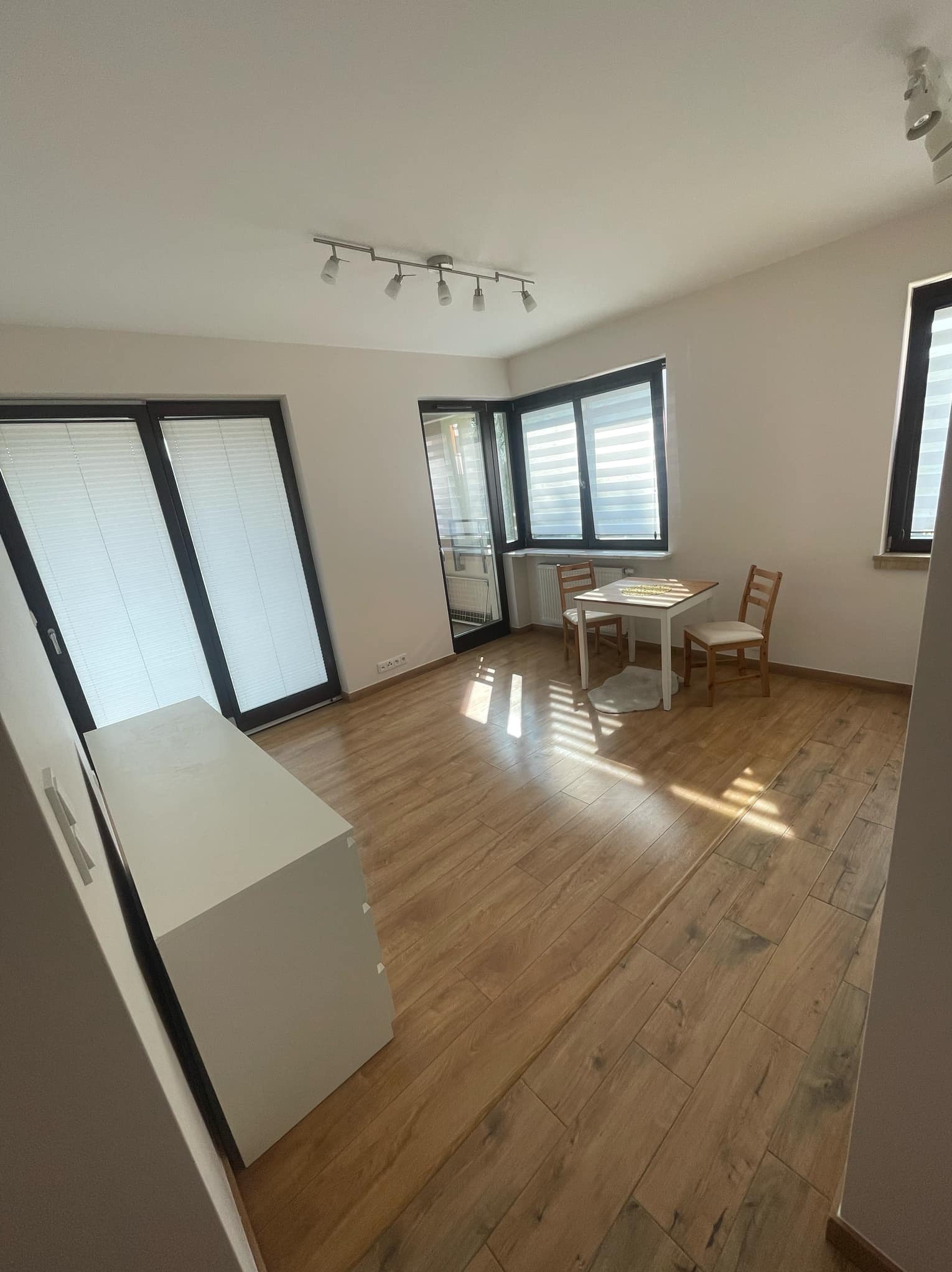 Jasne wnętrze pokoju z drewnopodobną podłogą, białymi ścianami, oknami z żaluzjami i stołem z krzesłami w tle, oświetlone punktowymi lampami sufitowymi.