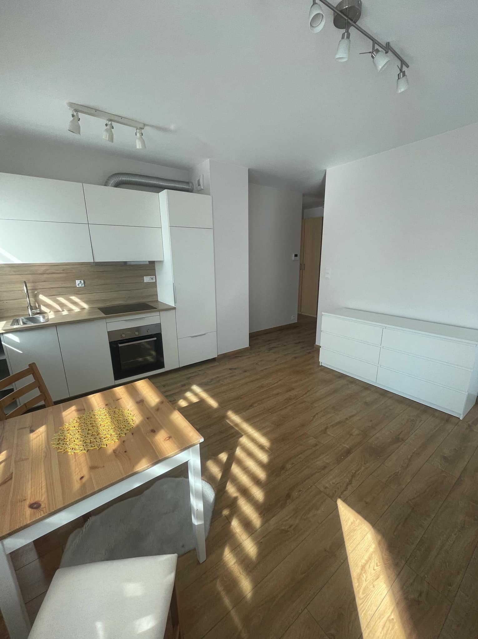 Jasne wnętrze mieszkania po remoncie: panele podłogowe, biała zabudowa kuchenna, stół z dekoracją w kształcie gwiazdy, komoda i oświetlenie punktowe.