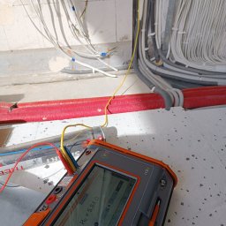 INSTALACJE ELEKTRYCZNE Łukasz Hibszer - Miernik rezystancji uziemienia, model Sonel MRU-536, z podłączonymi przewodami pomiarowymi, wyświetlający wynik pomiaru Re=5,84 Ω, na tle surowych ścian i wiązek kabli elektrycznych w peszlach...