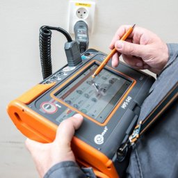 INSTALACJE ELEKTRYCZNE Łukasz Hibszer - Elektryk używa miernika MPI-540 z pomarańczowymi akcentami i rysikiem do testowania gniazdka elektrycznego z żółtą etykietą RB1/F27 na białej ścianie.