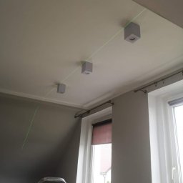 INSTALACJE ELEKTRYCZNE Łukasz Hibszer - Montaż trzech kwadratowych lamp sufitowych z użyciem lasera krzyżowego do poziomowania, widoczna drabina i okno w tle.