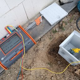 INSTALACJE ELEKTRYCZNE Łukasz Hibszer - Pomiar rezystancji uziemienia w Idzikowicach przy użyciu miernika Sonel, widoczna skrzynka przyłączeniowa w wykopie, zestaw kluczy imbusowych i wiertarka obok.