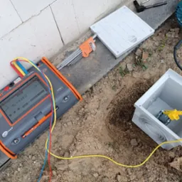 Pomiar rezystancji uziemienia w Idzikowicach przy użyciu miernika Sonel, widoczna skrzynka przyłączeniowa w wykopie, zestaw kluczy imbusowych i wiertarka obok.