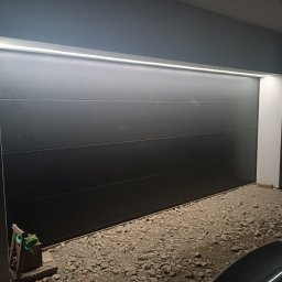 INSTALACJE ELEKTRYCZNE Łukasz Hibszer - Zamknięta, segmentowa brama garażowa w kolorze antracytowym z oświetleniem LED nadprożowym, widoczny fragment podjazdu wysypanego żwirem i fragmenty zaparkowanych samochodów.