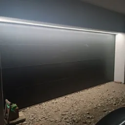 Zamknięta, segmentowa brama garażowa w kolorze antracytowym z oświetleniem LED nadprożowym, widoczny fragment podjazdu wysypanego żwirem i fragmenty zaparkowanych samochodów.