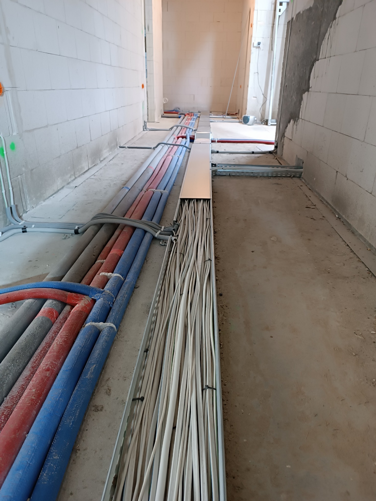 Wiązki kabli elektrycznych i rur instalacyjnych ułożone w korytach i na podłodze w surowym stanie budynku, widoczne ślady kurzu budowlanego.