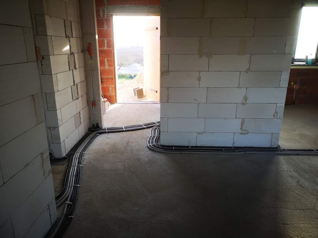 Wnętrze w trakcie budowy: widoczne ściany z białego bloczka, świeża wylewka podłogowa oraz poprowadzone wiązki przewodów elektrycznych wzdłuż ścian i przez otwór drzwiowy.