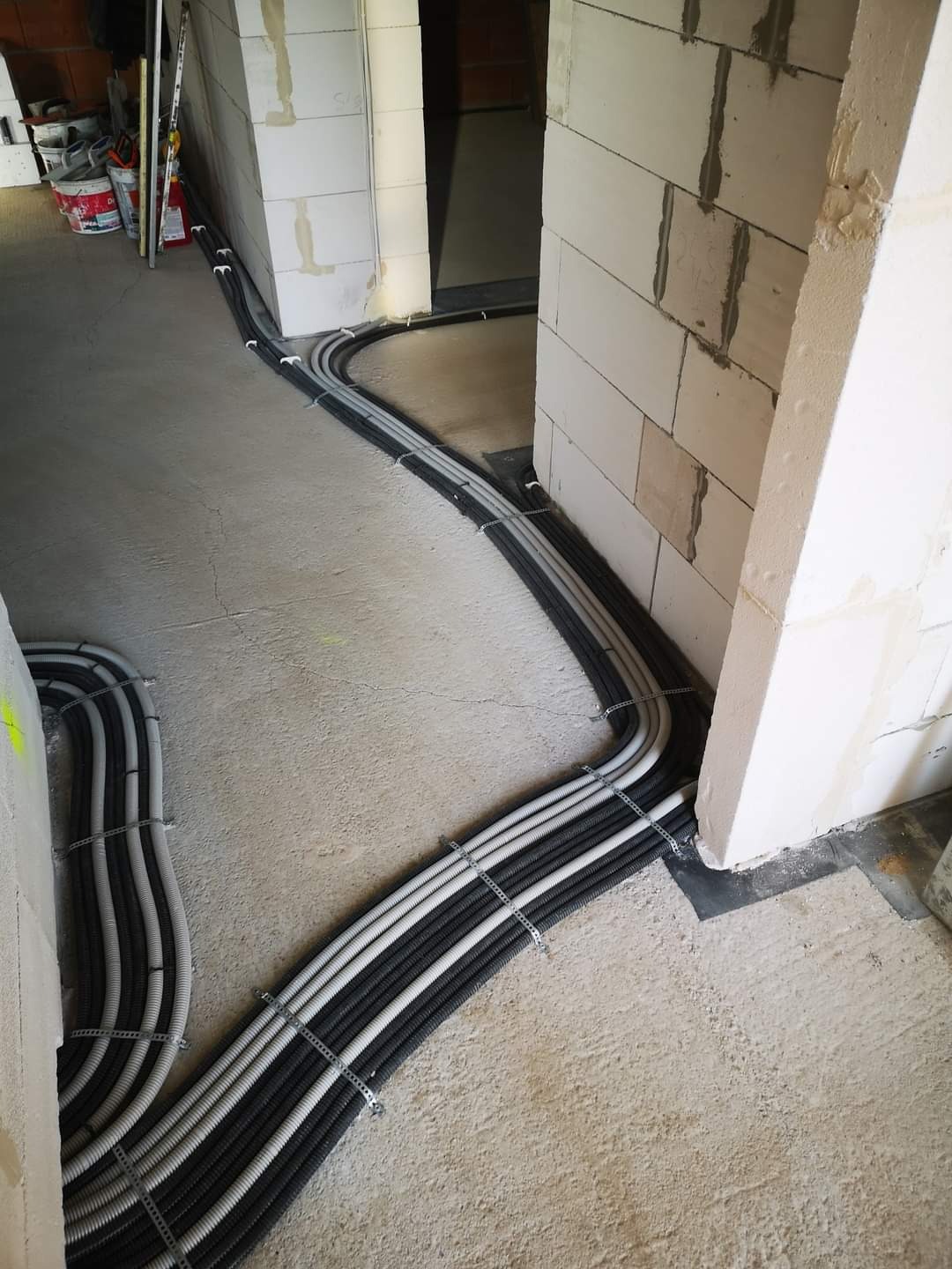 Równolegle ułożone rury instalacyjne, czarne i szare, przymocowane metalowymi uchwytami do surowego, betonowego podłoża w nowym budynku z białych bloczków.