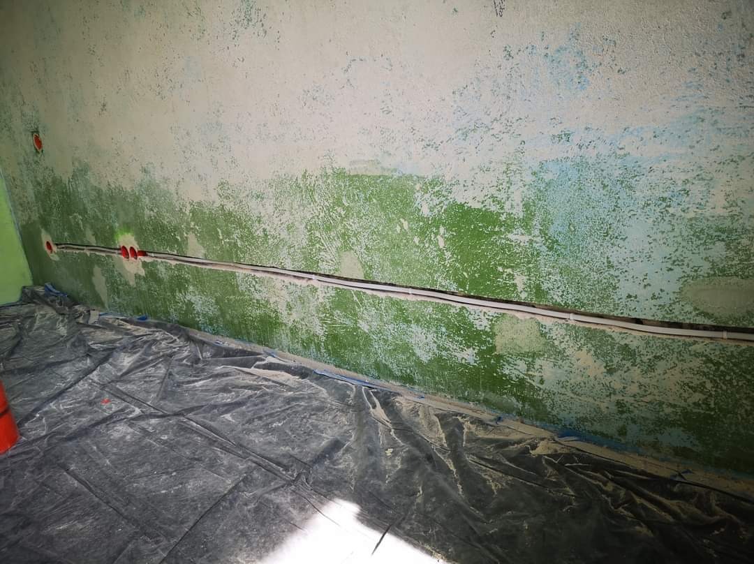 Biały kabel elektryczny ułożony w wyżłobieniu w ścianie o nierównej, zielono-białej powierzchni. Widoczne czerwone puszki instalacyjne. Podłoga zabezpieczona folią malarską.