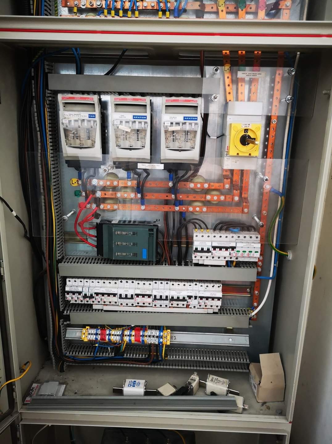 Otwarta szafa elektryczna z zamontowanymi bezpiecznikami, wyłącznikami nadprądowymi, sterownikiem PLC i okablowaniem, widoczne elementy miedziane i aluminiowe.