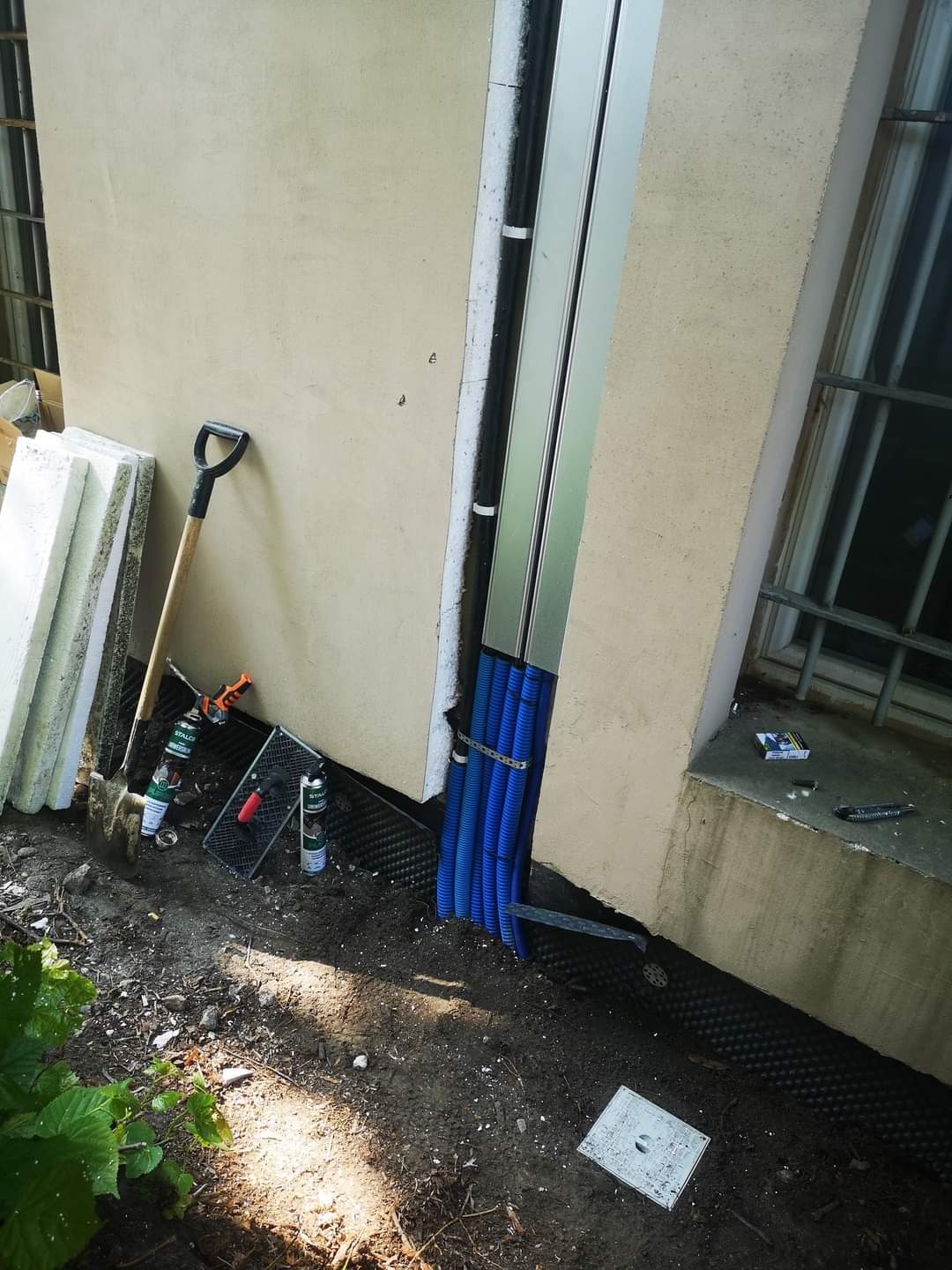 Przygotowanie do instalacji elektrycznej zewnętrznej: niebieskie rury karbowane, puszka elektryczna w ziemi, narzędzia (szpadel, pistolet do pianki, poziomica) przy ścianie budynku z oknem.