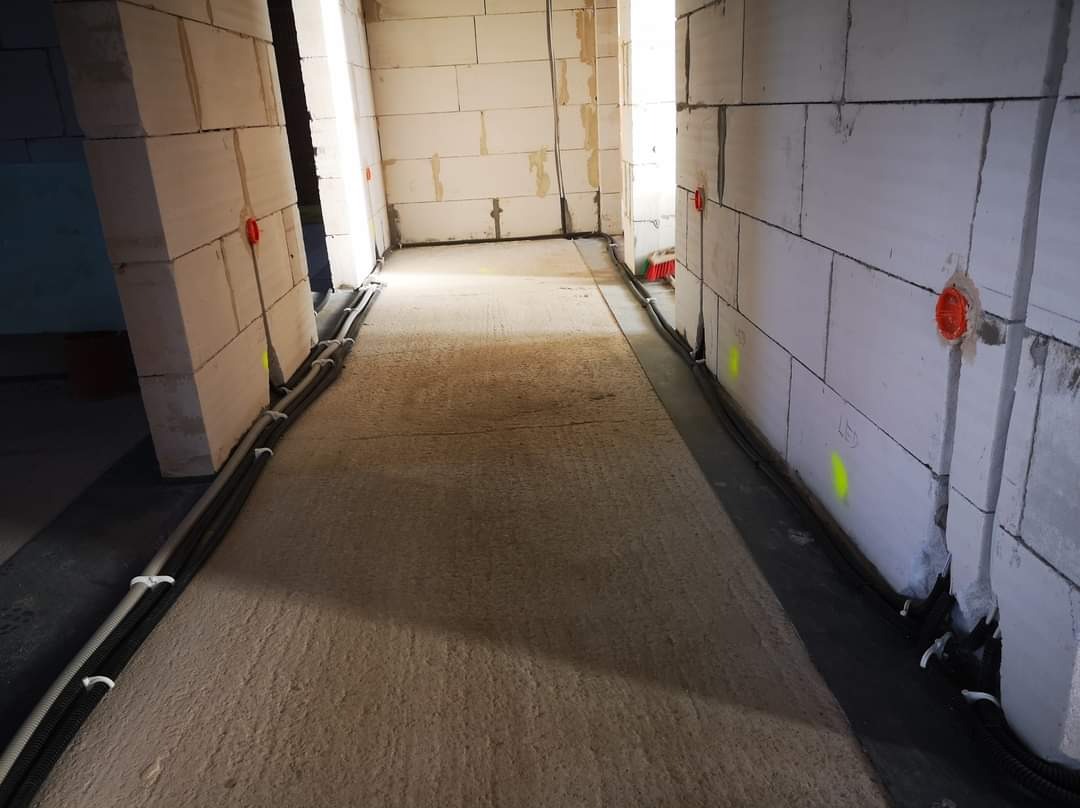 Wnętrze w stanie surowym z widocznymi przewodami elektrycznymi w peszlach, przymocowanymi do ścian z betonu komórkowego za pomocą uchwytów, oraz pomarańczowymi puszkami instalacyjnymi.