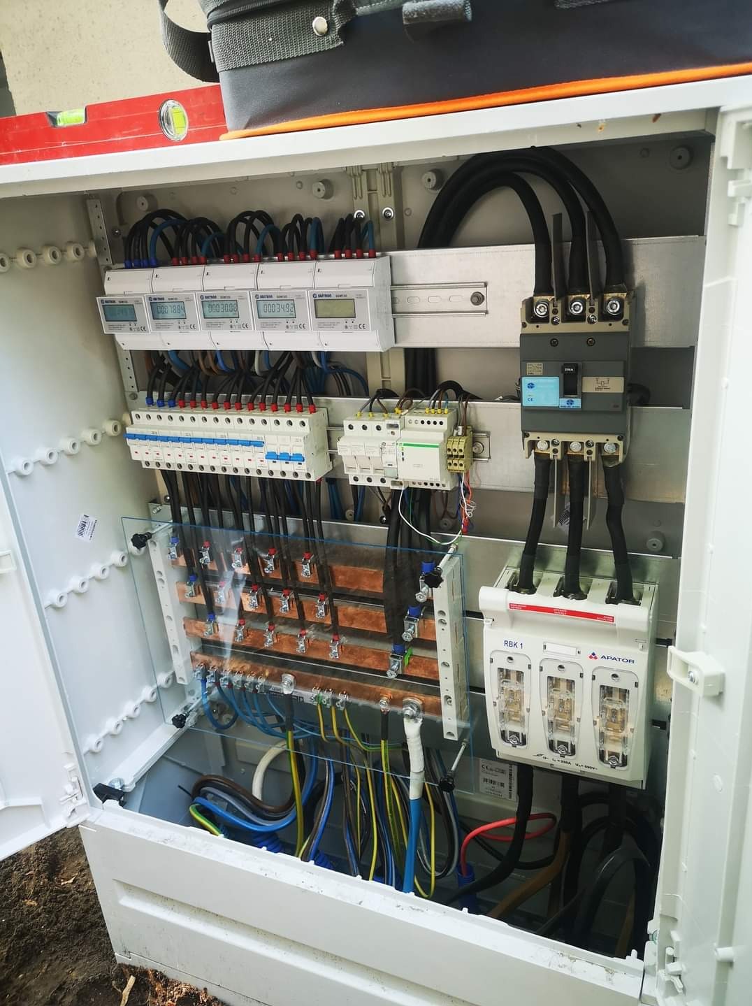Otwarta skrzynka elektryczna z zamontowanymi licznikami, bezpiecznikami i rozdzielnicą, widoczny poziomica na górnej krawędzi obudowy.