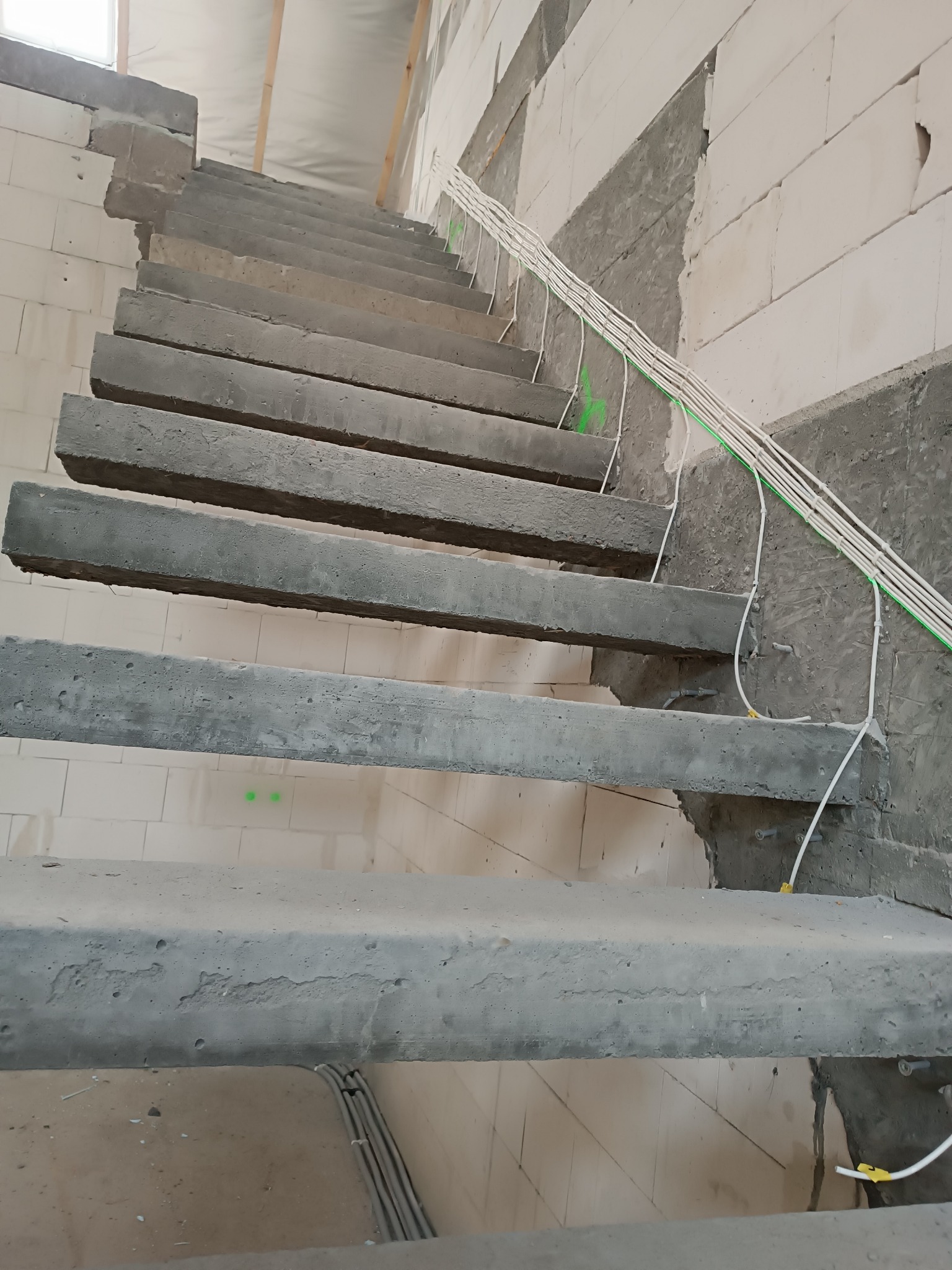 Surowe betonowe schody w trakcie budowy, z widocznymi przewodami elektrycznymi przymocowanymi do ściany, surowy stan wykończenia wnętrza z bloczków i częściowo otynkowanej ściany.
