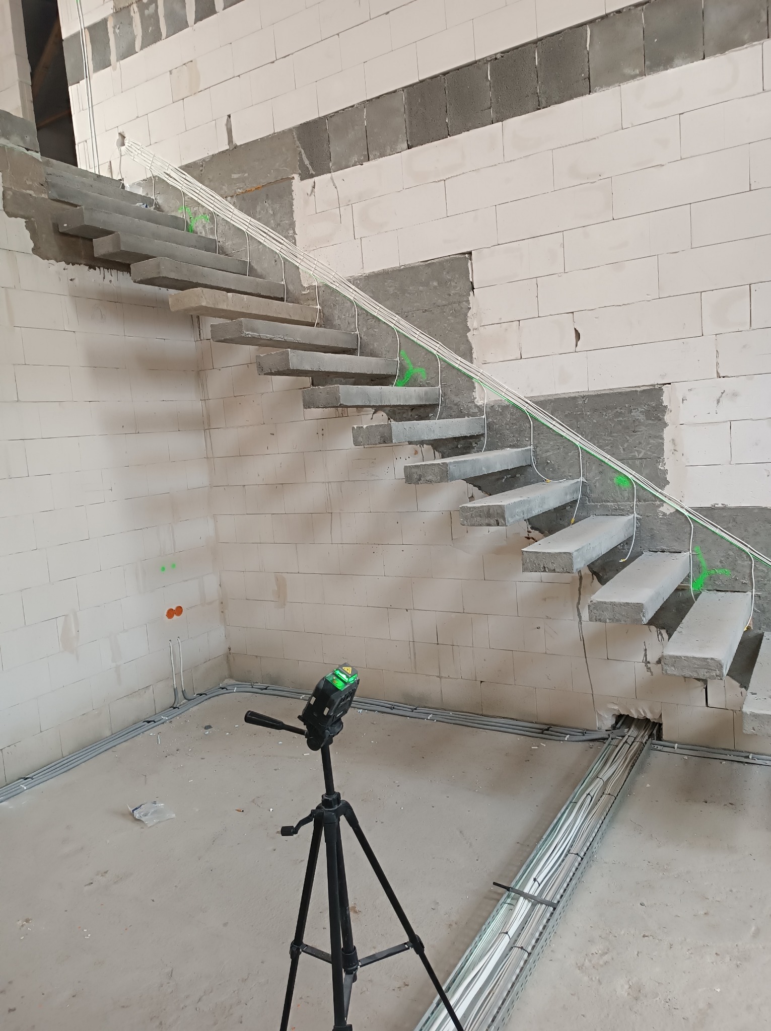 Surowe betonowe schody wiszące w trakcie montażu instalacji elektrycznej, widoczny laser krzyżowy na statywie, kable prowadzone w korytkach i po ścianie z betonu komórkowego.