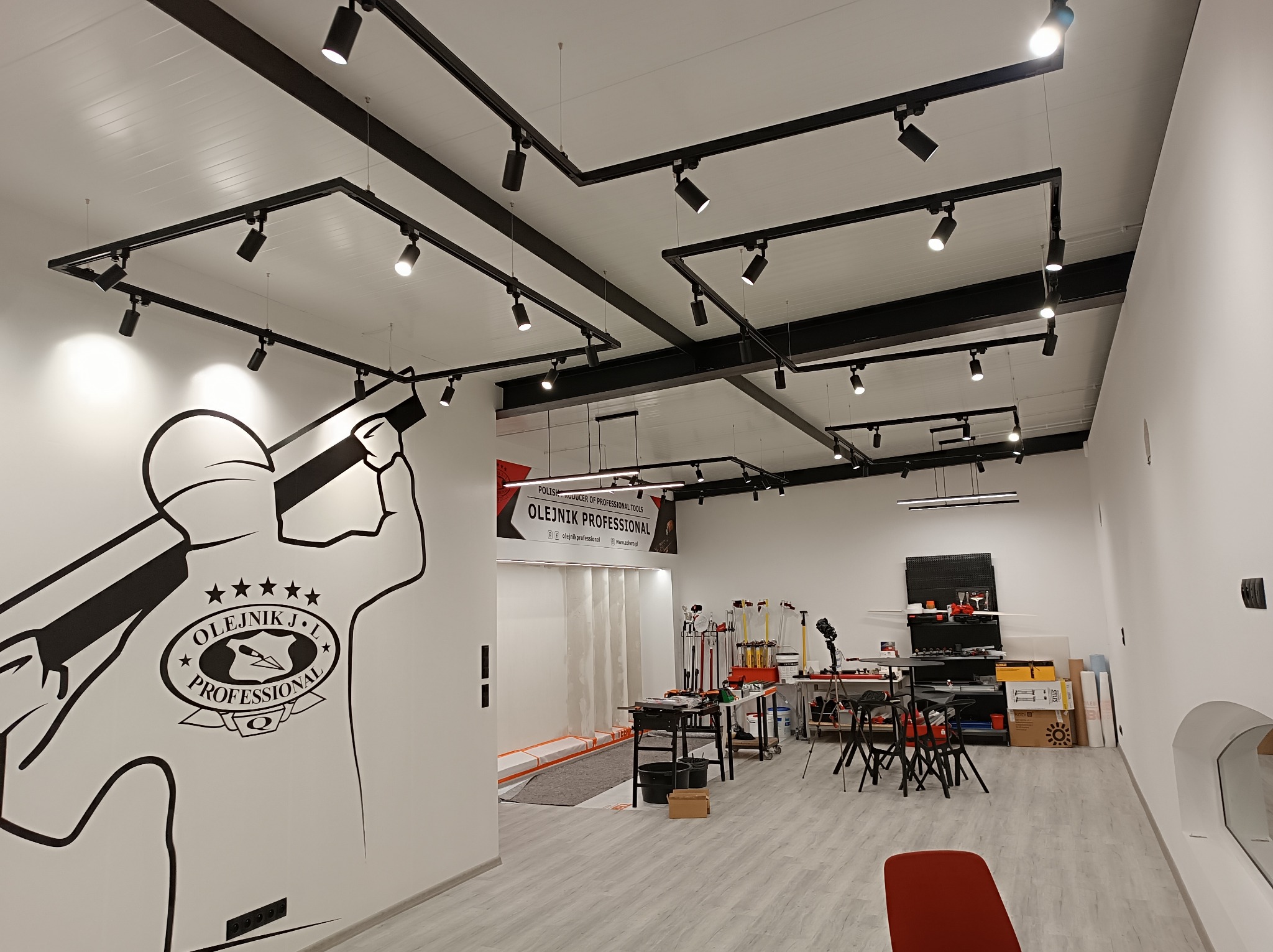 Wnętrze jasnego warsztatu lub pracowni z systemem szynoprzewodów oświetleniowych na suficie, narzędzia i stanowiska pracy w tle, na ścianie grafika z logotypem.