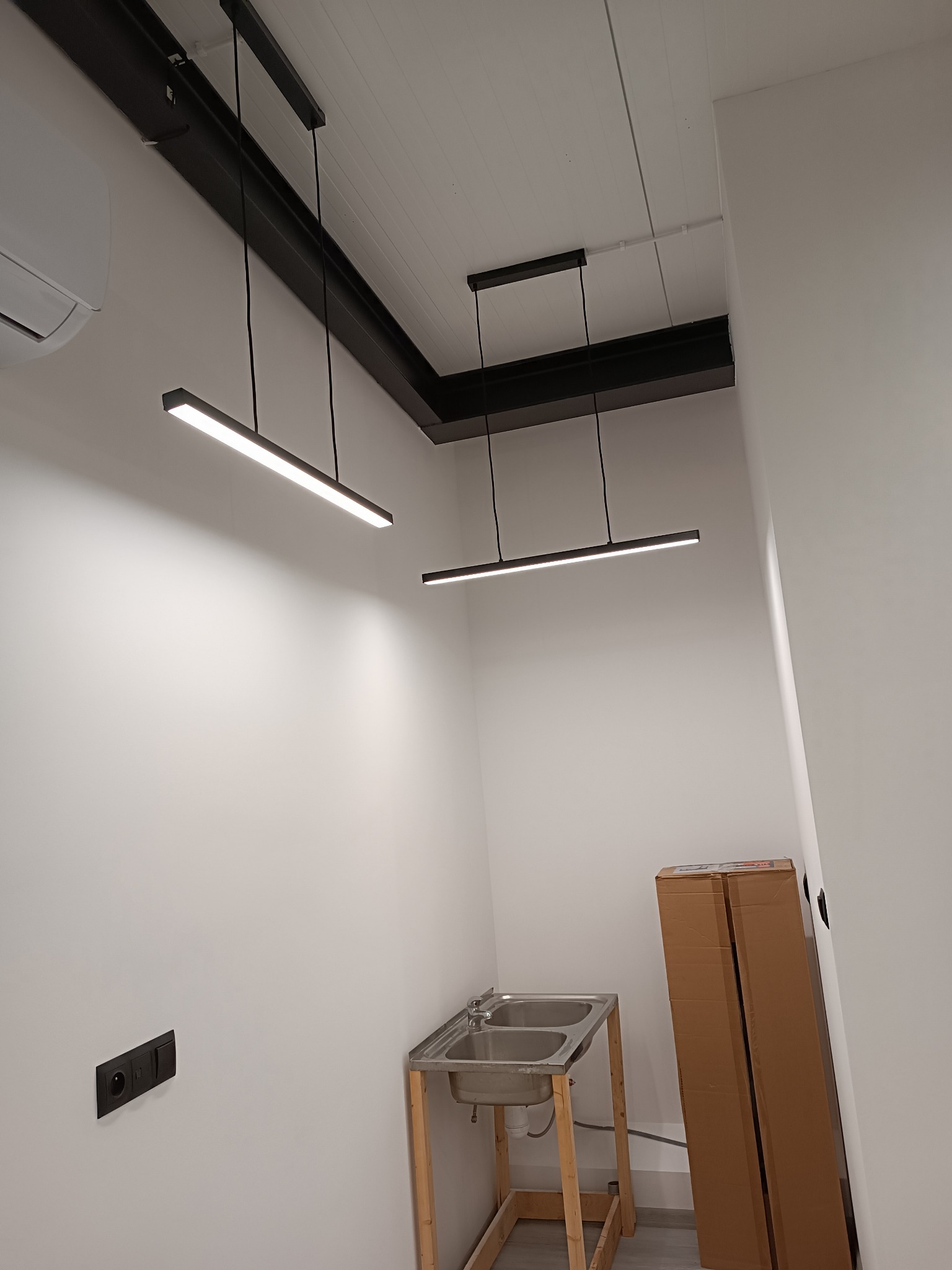 Dwa nowoczesne, liniowe źródła światła LED zawieszone na czarnych linkach, zamontowane na czarnej szynie sufitowej w pomieszczeniu z podwójnym zlewem na drewnianej konstrukcji i klimatyzatorem...