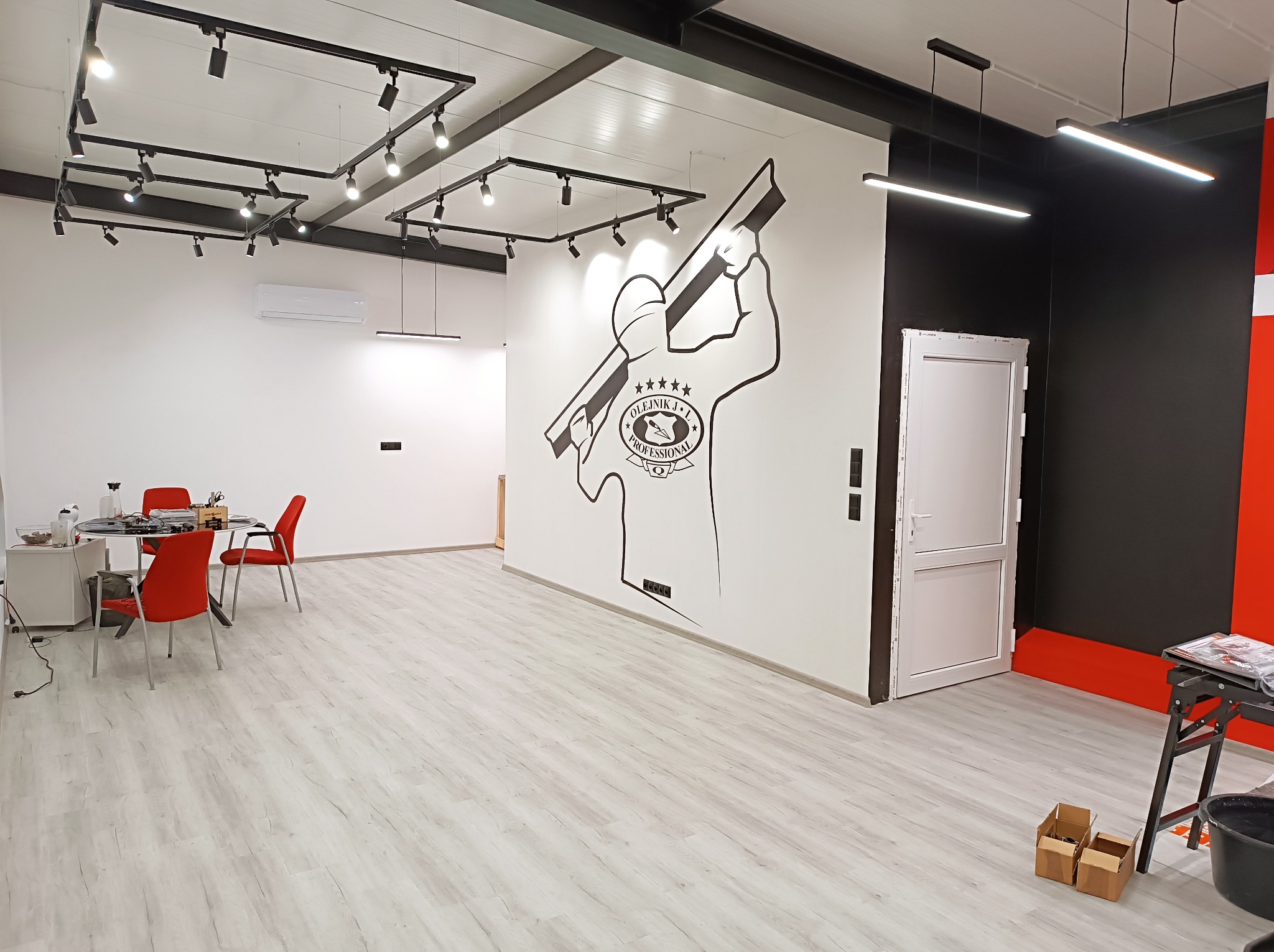 Wnętrze biura firmy z branży elektrycznej z nowoczesnym oświetleniem, białymi ścianami i podłogą imitującą drewno, z dużym logo firmy na ścianie przedstawiającym elektryka z narzędziem w dłoni.