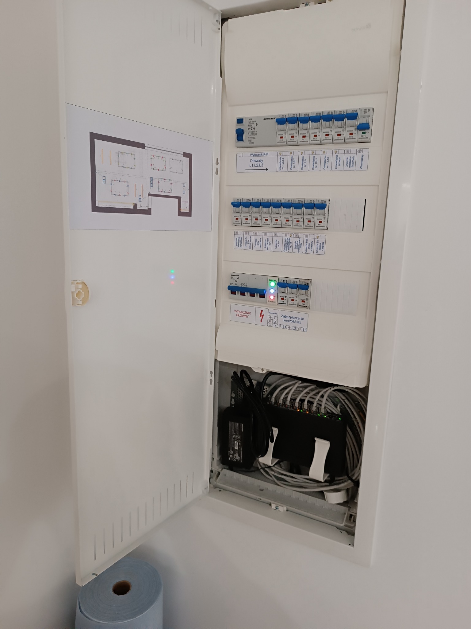 Otwarta szafka rozdzielcza z bezpiecznikami, wyłącznikami i przewodami. Na drzwiach szafki naklejony schemat instalacji elektrycznej mieszkania. Widoczne diody LED sygnalizujące pracę urządzeń.