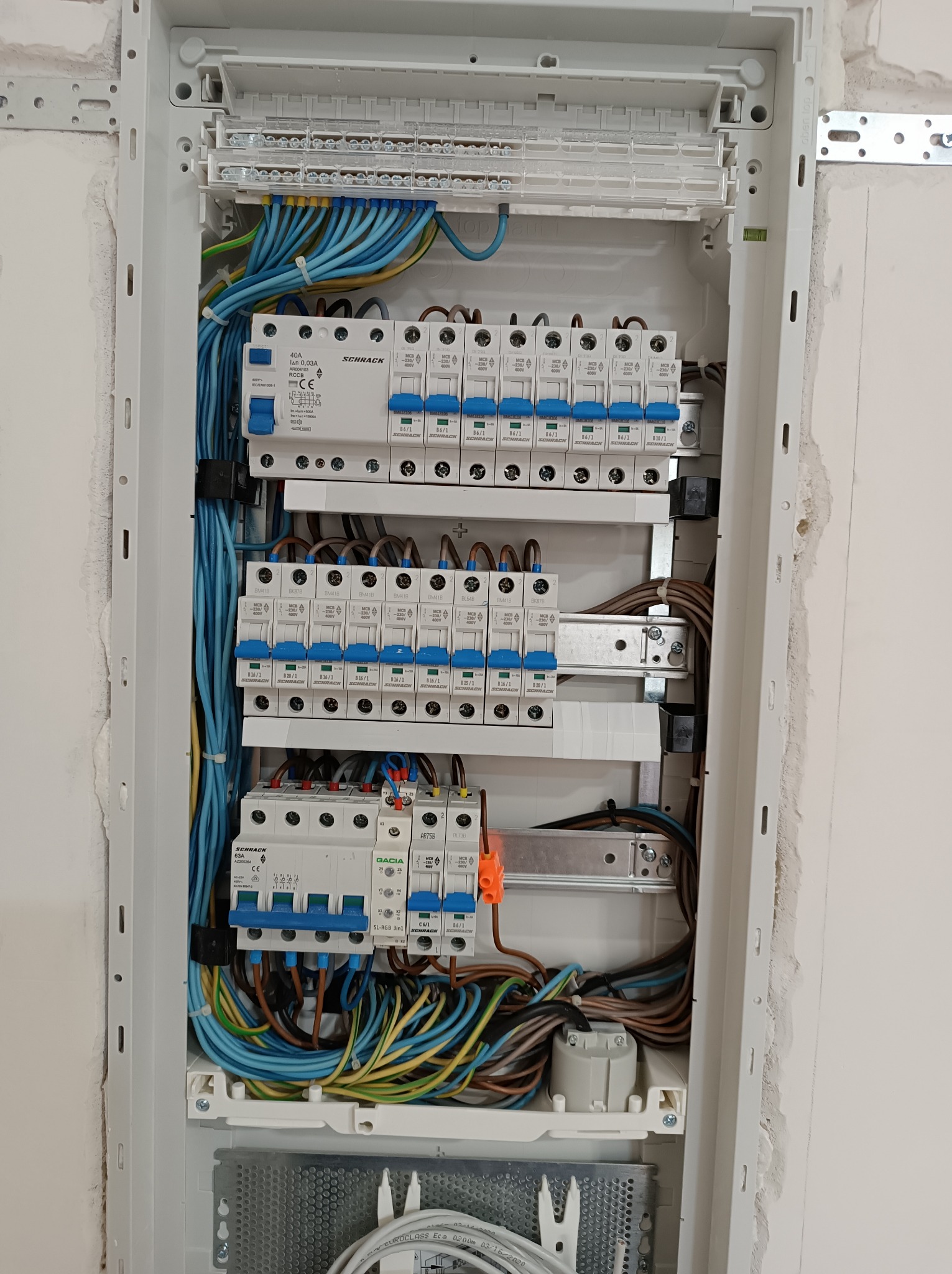 Rozdzielnica elektryczna w białej obudowie, widoczne liczne bezpieczniki Schrack i Gacia, okablowanie w kolorach niebieskim, brązowym, żółto-zielonym, zamontowana na ścianie z surowym tynkiem.