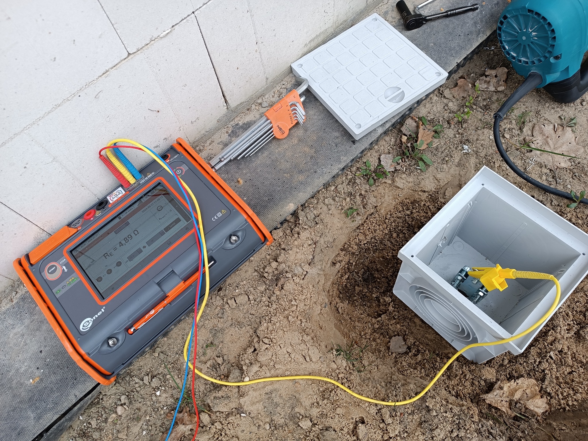 Pomiar rezystancji uziemienia w Idzikowicach przy użyciu miernika Sonel, widoczna skrzynka przyłączeniowa w wykopie, zestaw kluczy imbusowych i wiertarka obok.