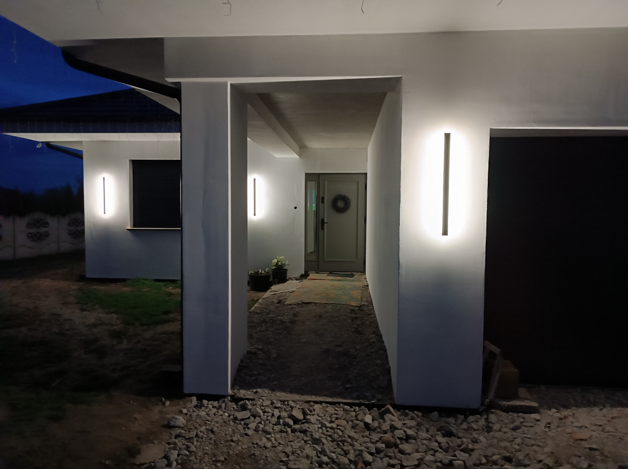 Nowoczesny dom z białymi ścianami i pionowymi lampami LED na elewacji, widok na wejście z dekoracyjnym wieńcem na drzwiach, teren wokół domu w trakcie prac wykończeniowych.