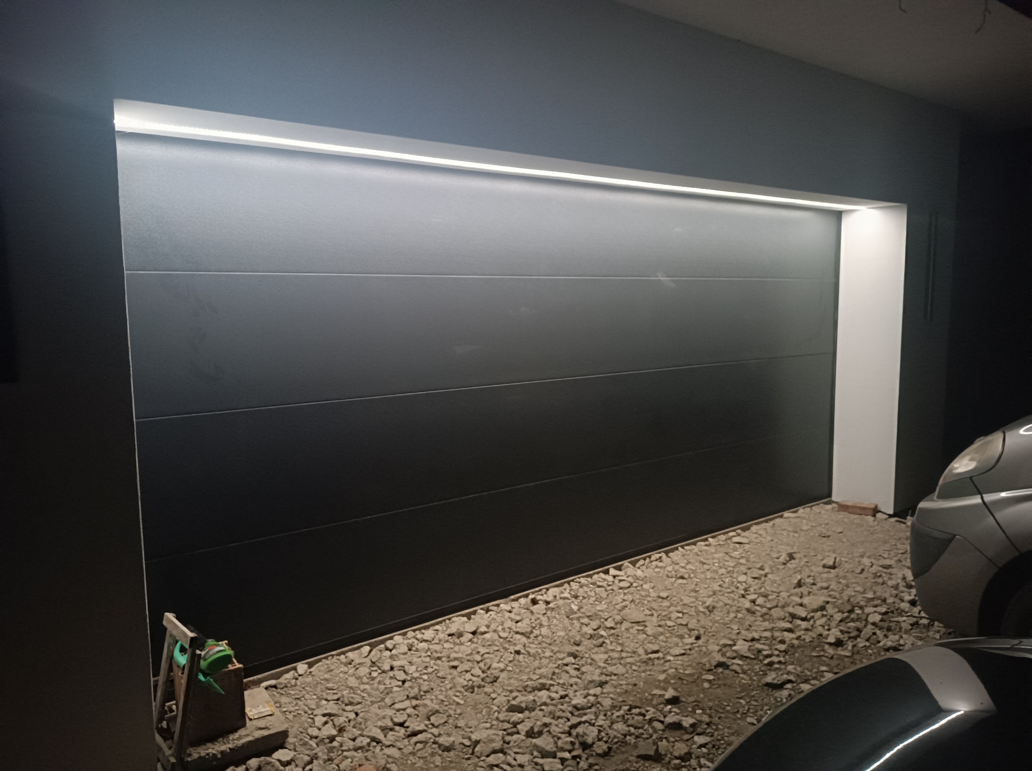 Zamknięta, segmentowa brama garażowa w kolorze antracytowym z oświetleniem LED nadprożowym, widoczny fragment podjazdu wysypanego żwirem i fragmenty zaparkowanych samochodów.