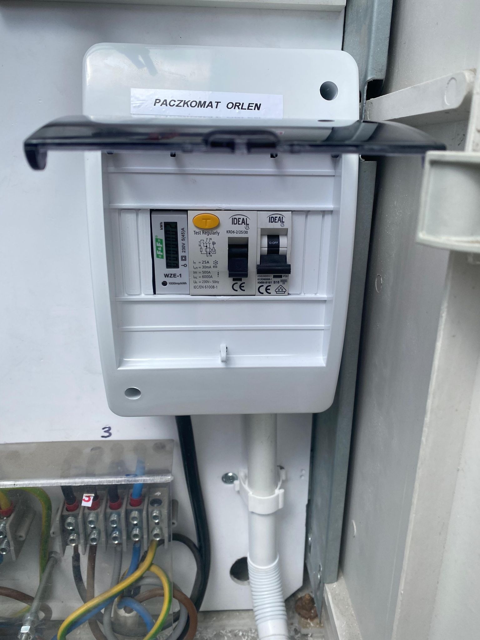 Otwarta skrzynka elektryczna z widocznymi bezpiecznikami IDEAL, licznikiem energii WZE-1 oraz naklejką 'PACZKOMAT ORLEN' na górnej klapie.