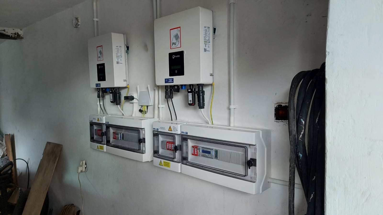Dwa inwertery fotowoltaiczne FoxESS zamontowane na białej ścianie, poniżej dwie skrzynki rozdzielcze z zabezpieczeniami, widoczny również kabel koncentryczny i fragment nieuporządkowanego otoczenia.