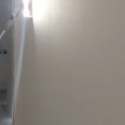 Świeżo wyszpachlowana ściana oświetlona przez otwór okienny z widocznym fragmentem wałka malarskiego i surowym wykończeniem ościeżnicy.