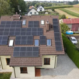 Widok z góry na dom z brązowym dachem pokrytym panelami słonecznymi, w tle zabudowa wiejska i pola uprawne.