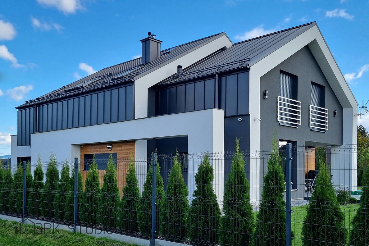 Nowoczesny dom jednorodzinny z szarą elewacją, czarnym dachem i metalowym ogrodzeniem obsadzonym tujami, widok z zewnątrz w słoneczny dzień.