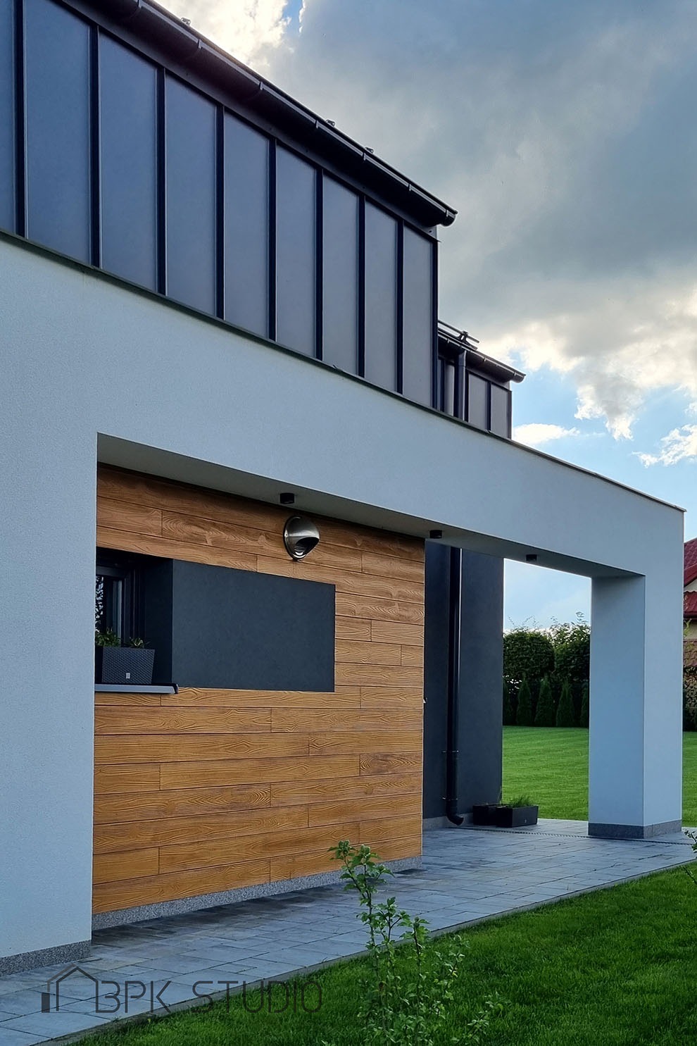 Nowoczesny dom jednorodzinny z elewacją w odcieniach bieli, drewna i grafitu, z widocznym oświetleniem zewnętrznym i fragmentem zadbanego trawnika.