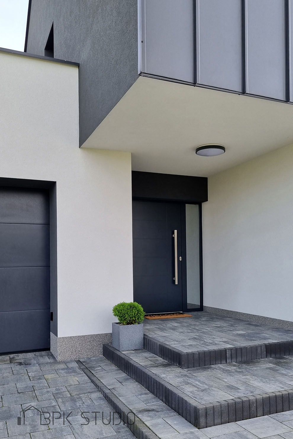 Nowoczesne wejście do domu z ciemnymi drzwiami i szarymi schodami z kostki brukowej, ozdobione małą kulistą rośliną w szarej donicy.