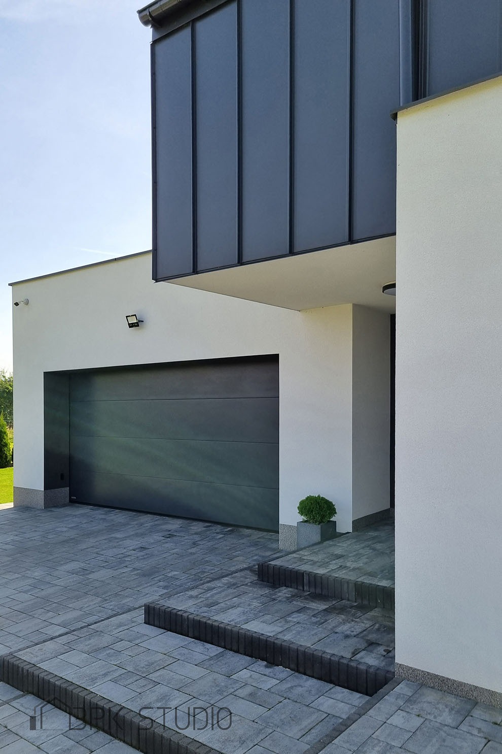 Nowoczesny dom jednorodzinny z grafitową bramą garażową, elewacją w odcieniach bieli i szarości oraz brukowanym podjazdem z kamiennymi schodami.